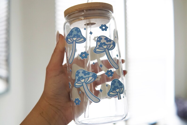 May include: A clear glass tumbler with a bamboo lid and a glass straw. The exterior of the glass features a repeating pattern of blue mushrooms and small flowers, accented with tiny four-pointed star shapes scattered across the surface.