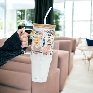 May include: A clear glass tumbler with a white handle and a bamboo lid. The tumbler features floral designs and inspirational text. A white straw is inserted into the tumbler. The tumbler is held by a person.