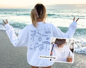 Op de afbeelding: Witte sweatshirt met blauwe illustraties en tekst. Het ontwerp bevat een cocktail, badpak, parasol en de tekst "With love from The Hamptons". Een kleinere afbeelding toont het ontwerp aan de voorkant. De persoon staat op een strand.