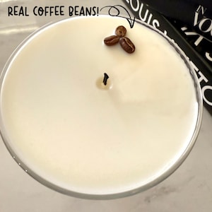 May include: A white candle in a clear glass container with a black wick and a small cluster of coffee beans on top. The text "REAL COFFEE BEANS!" is written in black on a white background.