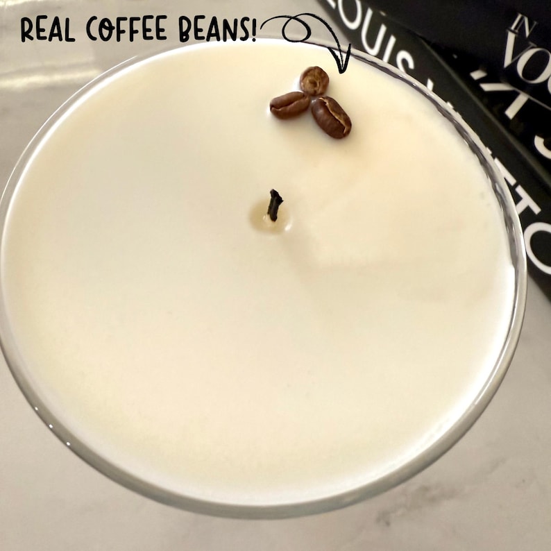 May include: A white candle in a clear glass container with a black wick and a small cluster of coffee beans on top. The text "REAL COFFEE BEANS!" is written in black on a white background.
