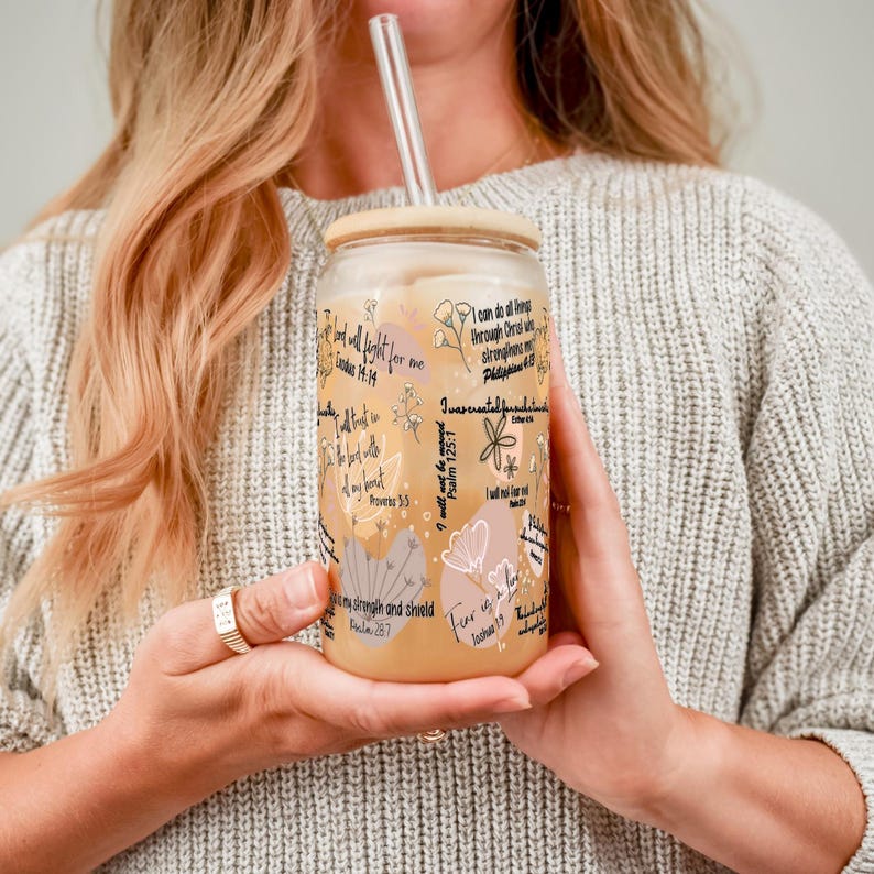 May include: A clear glass tumbler with a bamboo lid and a glass straw, filled with a light-colored beverage. The tumbler is decorated with floral designs and inspirational text in black. The tumbler is held in both hands.