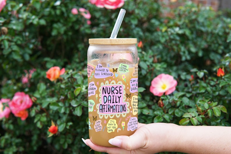 K&ouml;nnte beinhalten: Ein klares Glas mit Bambusdeckel und einem durchsichtigen Strohhalm, gef&uuml;llt mit einer hellbraunen Fl&uuml;ssigkeit. Der Becher ist mit bunten Aufklebern mit der Aufschrift "Nurse Daily Affirmations" und anderen positiven Affirmationen verziert. Der Hintergrund ist ein &uuml;ppiger gr&uuml;ner Busch mit rosa und orangefarbenen Bl&uuml;ten.