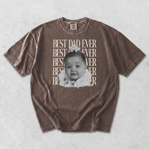 Puede incluir: Camiseta marr&oacute;n con el texto "BEST DAD EVER" apilado verticalmente a la izquierda y horizontalmente a la derecha. Una imagen en blanco y negro de un beb&eacute; est&aacute; en el centro. El beb&eacute; lleva un atuendo blanco y tiene una flor en el pelo. El nombre del beb&eacute;, "Tatiana", est&aacute; escrito debajo.