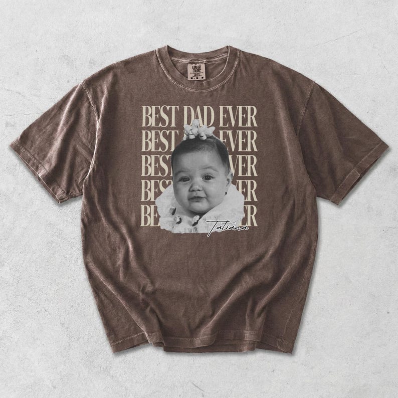 Puede incluir: Camiseta marr&oacute;n con el texto "BEST DAD EVER" apilado verticalmente a la izquierda y horizontalmente a la derecha. Una imagen en blanco y negro de un beb&eacute; est&aacute; en el centro. El beb&eacute; lleva un atuendo blanco y tiene una flor en el pelo. El nombre del beb&eacute;, "Tatiana", est&aacute; escrito debajo.