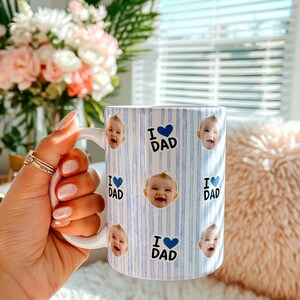 K&ouml;nnte beinhalten: Wei&szlig;er Keramikbecher mit blau-wei&szlig;em Streifenmuster. Der Becher zeigt Babygesichter und den Text "I ❤️ DAD" in Blau. Der Becher hat einen wei&szlig;en Henkel und ist f&uuml;r Hei&szlig;getr&auml;nke.