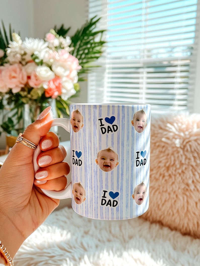 K&ouml;nnte beinhalten: Wei&szlig;er Keramikbecher mit blau-wei&szlig;em Streifenmuster. Der Becher zeigt Babygesichter und den Text "I ❤️ DAD" in Blau. Der Becher hat einen wei&szlig;en Henkel und ist f&uuml;r Hei&szlig;getr&auml;nke.