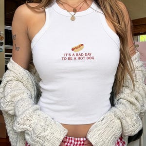 以下が含まれることがあります： 白いリブ編みのレーサーバックタンクトップ。ホットドッグの小さな刺繍と「IT'S A BAD DAY TO BE A HOT DOG」という赤い文字がデザインされています。クリーム色の厚手のニットカーディガンと、赤と白のギンガムチェック柄のパンツを合わせています。