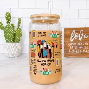 May include: A clear glass tumbler with a bamboo lid and a straw, filled with a light brown liquid. The tumbler is decorated with various graphics and text from the TV show "Friends", including the Central Perk logo and character illustrations.