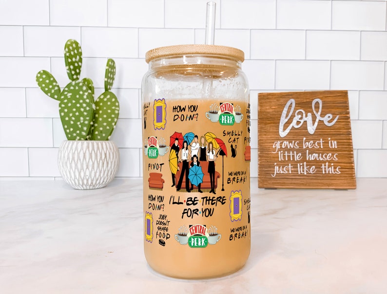 May include: A clear glass tumbler with a bamboo lid and a straw, filled with a light brown liquid. The tumbler is decorated with various graphics and text from the TV show "Friends", including the Central Perk logo and character illustrations.