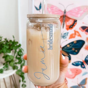 Puede incluir: Un vaso de cristal transparente con tapa de bamb&uacute; y pajita de cristal, lleno de una bebida helada de color beige. El vaso presenta el nombre Jamie escrito en una fuente cursiva azul, con la palabra bridesmaid en una fuente azul menor.