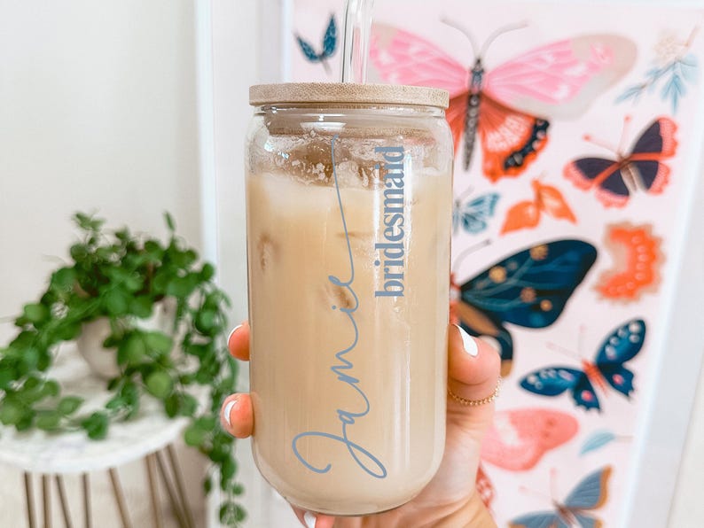 Puede incluir: Un vaso de cristal transparente con tapa de bamb&uacute; y pajita de cristal, lleno de una bebida helada de color beige. El vaso presenta el nombre Jamie escrito en una fuente cursiva azul, con la palabra bridesmaid en una fuente azul menor.