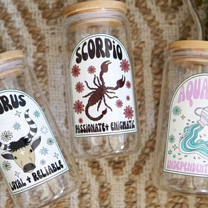 Pode incluir: Tr&ecirc;s frascos de vidro transparente com tampas de bambu, cada um com um signo do zod&iacute;aco diferente: Touro, Escorpi&atilde;o e Aqu&aacute;rio. Os frascos t&ecirc;m r&oacute;tulos coloridos com s&iacute;mbolos do zod&iacute;aco e tra&ccedil;os associados. O fundo &eacute; uma textura tecida.