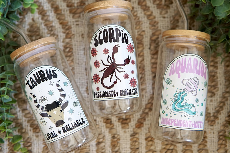 Pode incluir: Tr&ecirc;s frascos de vidro transparente com tampas de bambu, cada um com um signo do zod&iacute;aco diferente: Touro, Escorpi&atilde;o e Aqu&aacute;rio. Os frascos t&ecirc;m r&oacute;tulos coloridos com s&iacute;mbolos do zod&iacute;aco e tra&ccedil;os associados. O fundo &eacute; uma textura tecida.