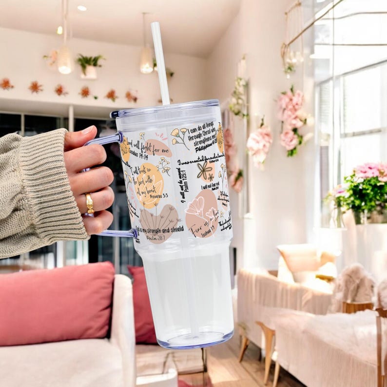 May include: Clear plastic tumbler with a white straw and a light blue handle. The tumbler is decorated with floral designs and inspirational text in black. The tumbler is designed to hold cold beverages.