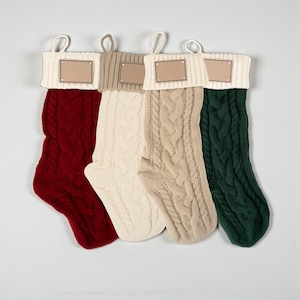 May include: Four cable-knit Christmas stockings in red, cream, tan, and green. Each stocking has a cream-colored cuff and a small, blank rectangular tag. The stockings are hung against a light gray background.