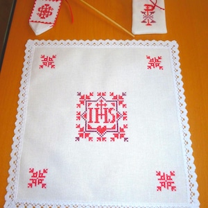 Könnte beinhalten: Ein weißes Leinentuch mit Spitzenborte und roter Kreuzstichstickerei. Das zentrale Design zeigt die Buchstaben "IHS" in einem Herzen und geometrischen Mustern. Eine kleine Fahne und ein Beutel mit passender Stickerei sind ebenfalls vorhanden.