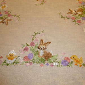 Könnte beinhalten: Eine beige Tischdecke mit gestickten Hasen, Küken und Blumen. Die Stickerei ist in Braun-, Gelb-, Rosa- und Grüntönen gehalten.