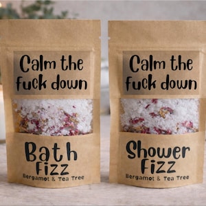 May include: Two brown paper pouches of bath and shower fizz. Each pouch has a clear window showing a mix of white granules and dried flowers. The text "Calm the fuck down" is above the window. One says "Bath Fizz" and the other "Shower Fizz".