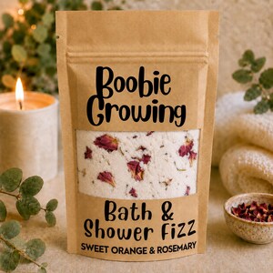 May include: A brown kraft paper pouch containing bath and shower fizz with visible dried rose petals. The label features the black text Boobie Growing Bath & Shower Fizz Sweet Orange & Rosemary. The pouch has a clear window showing the contents.