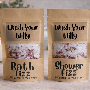 Peut inclure: Deux sachets en papier brun avec l'inscription "Wash Your Willy". L'un porte la mention "Bath Fizz", l'autre "Shower Fizz". Ils contiennent des sels de bain blancs avec des accents roses et dorés. "Bergamot & Tea Tree" est également visible.