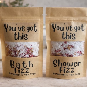 Puede incluir: Dos bolsas de papel marrón con efervescente para baño y ducha. Las bolsas tienen una ventana transparente que muestra sales de baño blancas con detalles en rojo y dorado. El texto en las bolsas dice "You've got this", "Bath Fizz" y "Shower Fizz".
