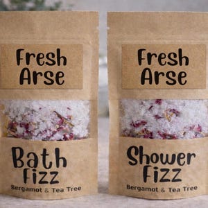 May include: Two brown paper pouches of bath products. The top label reads "Fresh Arse". The bottom labels read "Bath Fizz" and "Shower Fizz" with "Bergamot & Tea Tree" below. The pouches contain white bath salts with pink flower petals.