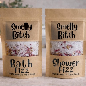 Puede incluir: Dos bolsas de papel kraft llenas de sales de baño y ducha. Las bolsas tienen una ventana transparente que muestra el producto, una mezcla de gránulos blancos y flores secas. El texto en las bolsas dice "Smelly Bitch" y "Bath Fizz" o "Shower Fizz".