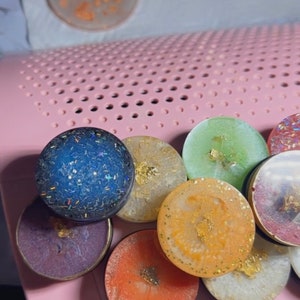 May include: A collection of colorful resin round ornaments with gold flakes and glitter. The ornaments are arranged in a semi-circle on a pink surface.