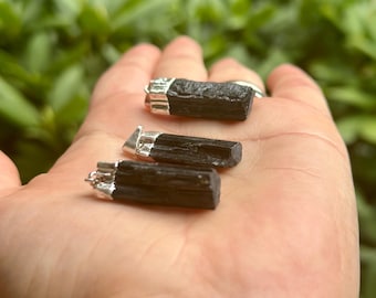 May include: Three black tourmaline pendants with silver-colored metal caps are displayed. The raw, natural crystals have a dark, textured appearance. The pendants are held in a hand against a blurred green background.