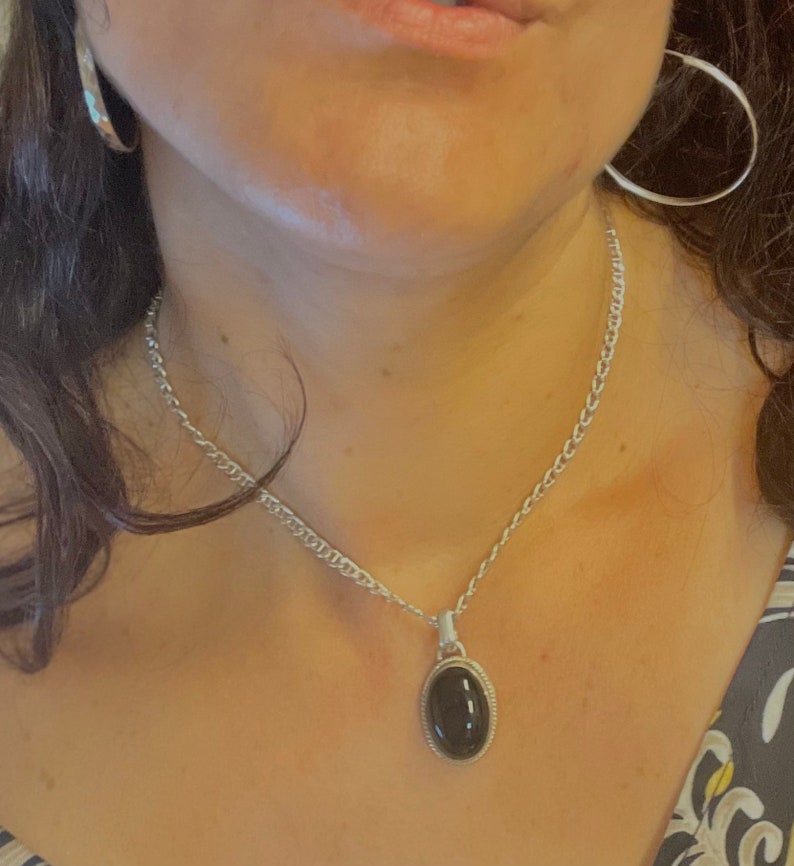 Op de afbeelding: Een zilveren ketting met een zwarte ovale hanger. De hanger heeft een zilveren rand.