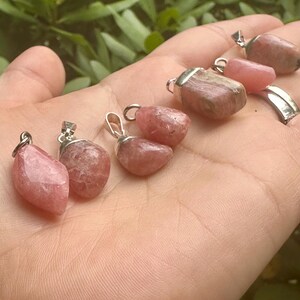 May include: A collection of pink gemstone pendants with silver-colored metal loops. The pendants vary in shape and shade, ranging from light to darker pink. The background is a blurred view of green foliage.