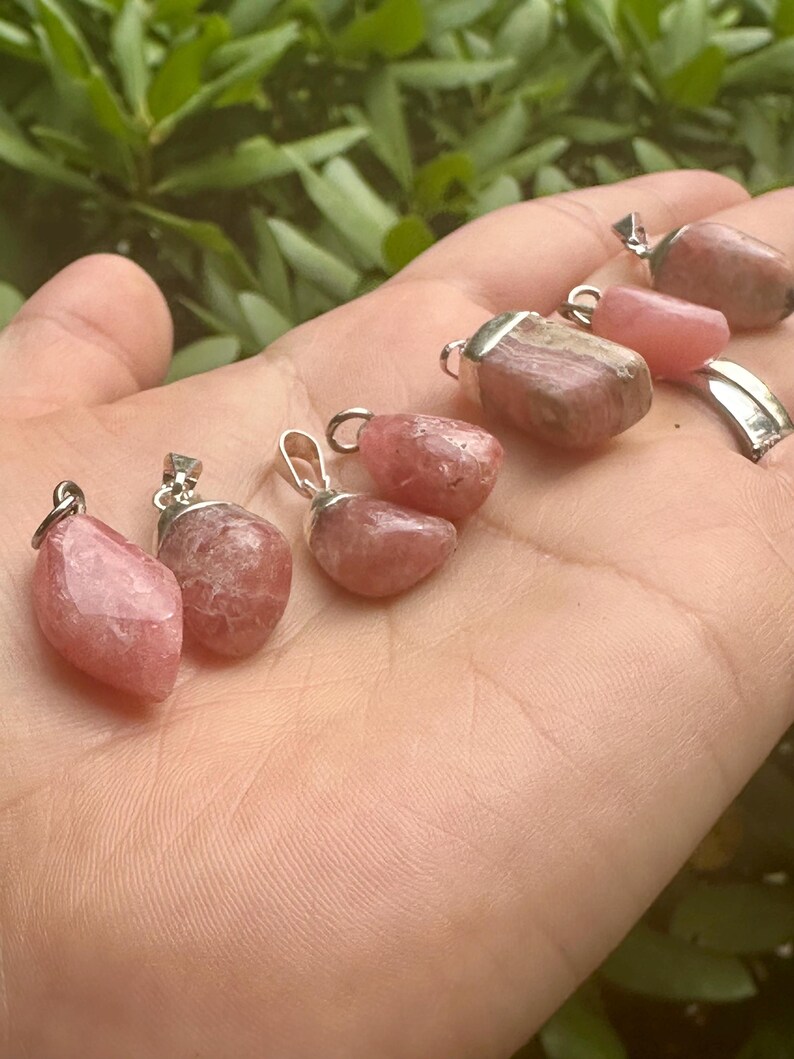 May include: A collection of pink gemstone pendants with silver-colored metal loops. The pendants vary in shape and shade, ranging from light to darker pink. The background is a blurred view of green foliage.