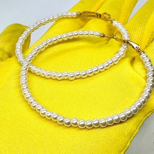 May include: A pair of large hoop earrings made of white faux pearls. The hoops are connected with a gold-toned clasp.