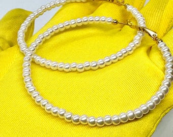 May include: A pair of large hoop earrings made of white faux pearls. The hoops are connected with a gold-toned clasp.