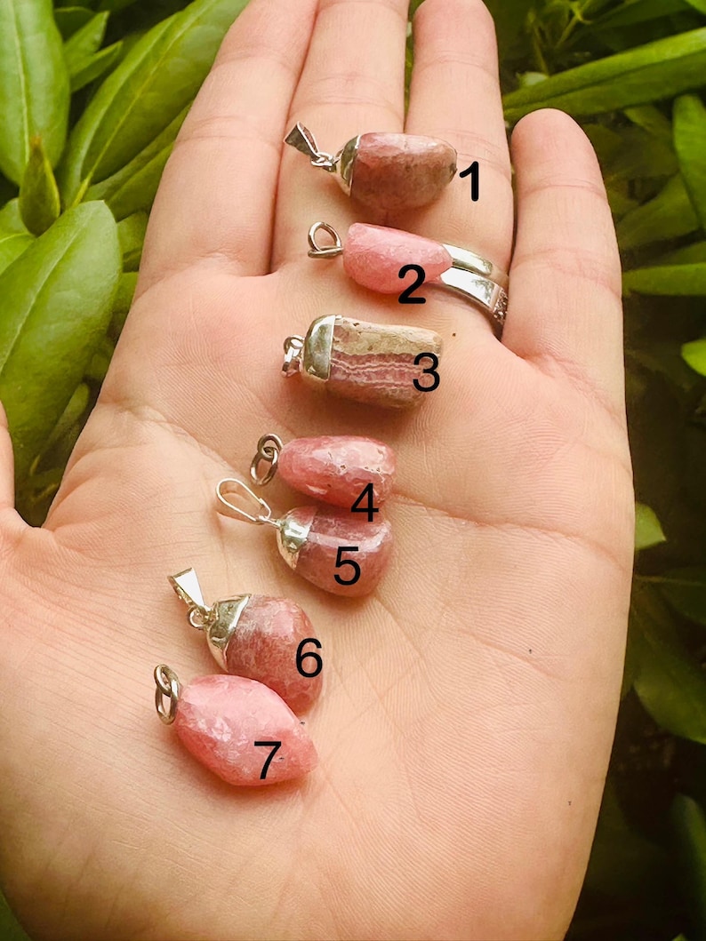 May include: Seven pink and tan gemstone pendants with silver-colored metal findings are displayed in a hand. The gemstones vary in shape and color, with some having a more solid pink hue and others showing a mix of pink and tan.