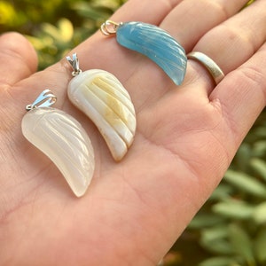 May include: Three carved gemstone pendants in the shape of wings. The pendants are made of white, light brown, and blue gemstones. The pendants are attached to silver chains.