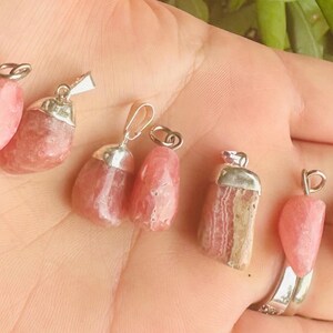 May include: A collection of pink gemstone pendants with silver-colored metal caps and loops. The pendants vary in shape and size, showcasing a range of pink hues. The background is a hand with green foliage.