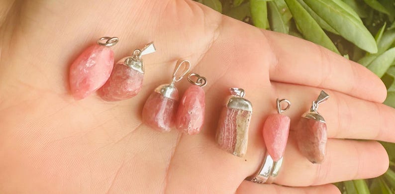 May include: A collection of pink gemstone pendants with silver-colored metal caps and loops. The pendants vary in shape and size, showcasing a range of pink hues. The background is a hand with green foliage.