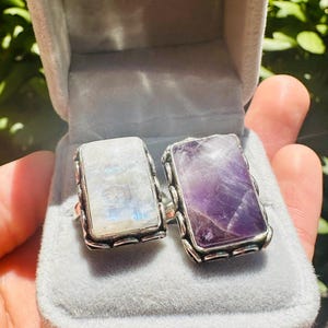 May include: Two gemstone rings in a gray velvet box. One ring features a rectangular, iridescent moonstone. The other ring has a rectangular, purple amethyst. Both rings have a silver-colored, textured setting.