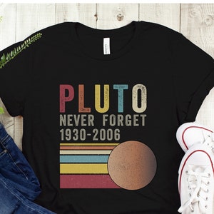 May include: Black t-shirt with a vintage-style graphic that says "Pluto Never Forget 1930-2006" with a colorful striped design and a brown circle representing Pluto.