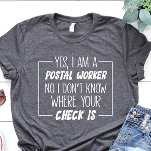 May include: A dark gray t-shirt with the text "YES, I AM A POSTAL WORKER NO I DON'T KNOW WHERE YOUR CHECK IS" printed in white.