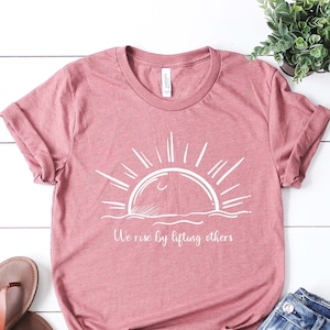 May include: A mauve t-shirt with a white graphic of a sun rising over the ocean. The text "We rise by lifting others" is printed below the graphic.