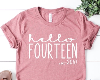 May include: A pink t-shirt with the text "hello fourteen est. 2010" in white lettering.
