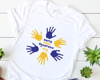 May include: White t-shirt with a blue and yellow handprint design that says "World Down Syndrome Day". The shirt is laid out on a white wooden surface with a pair of brown sandals and denim shorts.