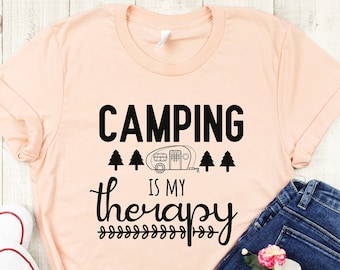 May include: A peach-colored t-shirt with the text "Camping is my therapy" in black. The text is surrounded by small trees and a camper icon. The shirt is laid out on a wooden surface with a pair of white sneakers and blue jeans.