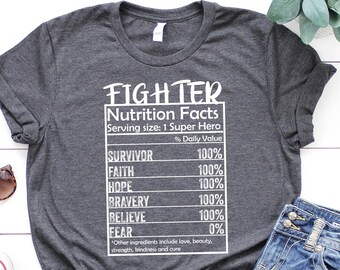 Op de afbeelding: Een donkergrijs T-shirt met witte tekst die "FIGHTER Nutrition Facts Serving size: 1 Super Hero % Daily Value SURVIVOR 100% FAITH 100% HOPE 100% BRAVERY 100% BELIEVE 100% FEAR 0% Other ingredients include love, beauty, strength, kindness and cure" luidt.