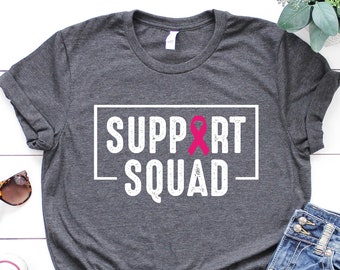 May include: A dark gray t-shirt with a white graphic that says "SUPPORT SQUAD" with a pink ribbon in the middle of the word "SUPPORT".