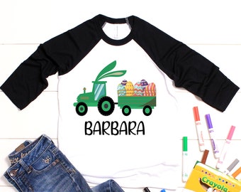 May include: A white and black baseball tee shirt with a green tractor carrying Easter eggs. The tractor has bunny ears and the text "BARBARA" is printed below the tractor.