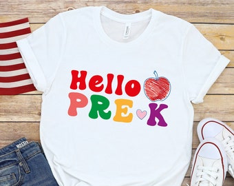 May include: White t-shirt with the text "Hello Pre-K" in colorful letters and a red apple drawing. The shirt is on a wooden surface with a pair of white sneakers and denim shorts.
